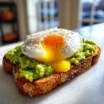 Close-up of perfectly mashed avocado toast topped with a poached egg with a runny yolk and cracked black pepper.