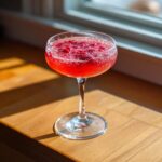 A vibrant raspberry honey lemon daiquiri served in a coupe glass, garnished with fresh raspberries, sitting on a sunlit wooden surface.