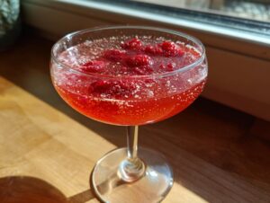 A vibrant raspberry honey lemon daiquiri garnished with fresh raspberries, sitting in a coupe glass on a sunlit wooden surface.