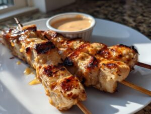 Three grilled sailor bailey bang bang chicken skewers served on a white plate with a side of dipping sauce.