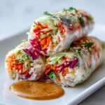 Three fresh sesame veggie spring rolls stacked on a white plate with a side of peanut dipping sauce.