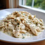 A close-up of shredded crockpot olive garden chicken coated in a creamy white sauce and sprinkled with green herbs.