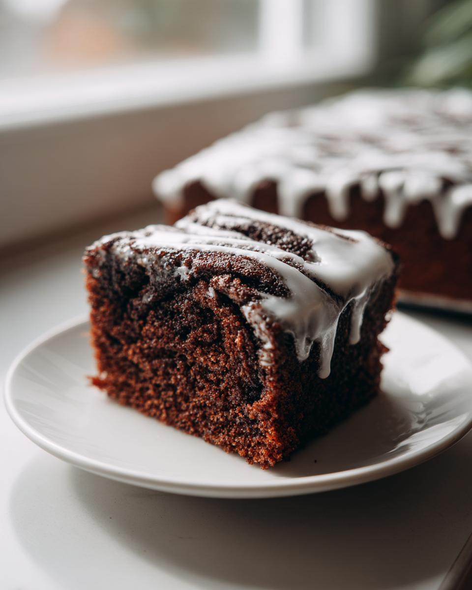 A rich, dark slice of gluten free cinnamon roll cake topped with thick white icing, served on a white plate.