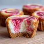 Close-up of a strawberry cheesecake cupcake with a bite taken out, showing creamy filling and graham cracker crust.
