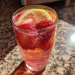 Close-up of a fizzy glass filled with strawberry lemonade punch, fresh strawberries, and a lemon slice.