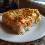 A square serving of sushi bake salmon resting on white rice, featuring flaky salmon and a golden, herbed, baked topping.