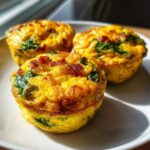 Close-up of three golden keto egg bites, baked with visible spinach and crispy bacon pieces, sitting on a white plate.