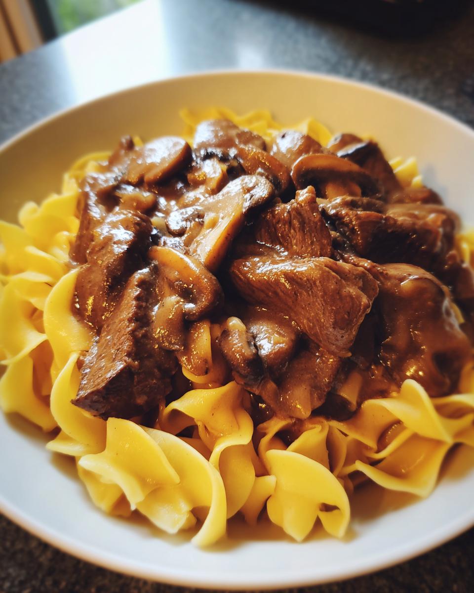 A white bowl filled with ultimate beef stroganoff, featuring tender beef chunks and mushrooms in a rich sauce served over wide egg noodles.