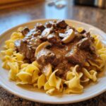 A close-up of ultimate beef stroganoff, featuring tender beef chunks and sliced mushrooms in a creamy brown sauce, served over wide egg noodles.