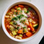 Overhead view of a bowl of rich vegetable minestrone soup, loaded with vegetables, pasta, and topped with grated Parmesan and parsley.