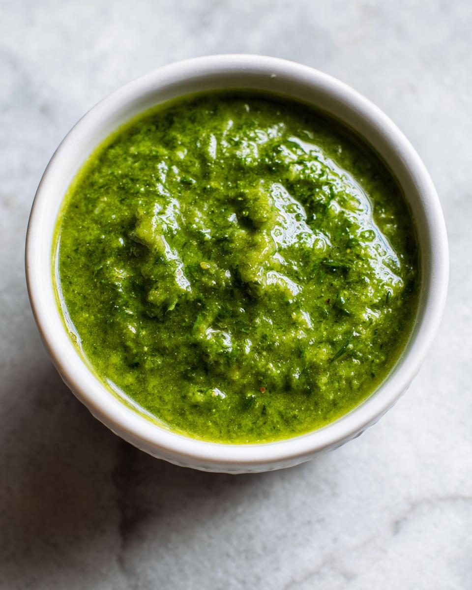Close-up overhead shot of thick, vibrant green enchilada sauce served in a small white bowl on a marble surface.