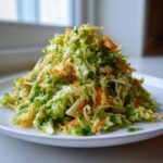 A tall mound of bright, freshly dressed mango slaw featuring shredded cabbage, carrots, and herbs on a white plate.