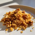 A close-up, brightly lit pile of golden walnut parm crumble mixed with whole walnut halves on a white plate.