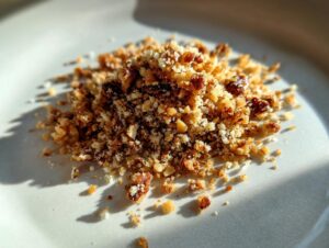A small pile of golden brown walnut parm crumble mixture sitting on a white plate under bright sunlight.