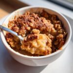 A spoonful of warm, gooey apple cinnamon crumble being lifted from a white ramekin.
