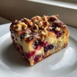 A square slice of Berry French toast casserole, loaded with red and blue berries, topped with walnuts on a white plate.