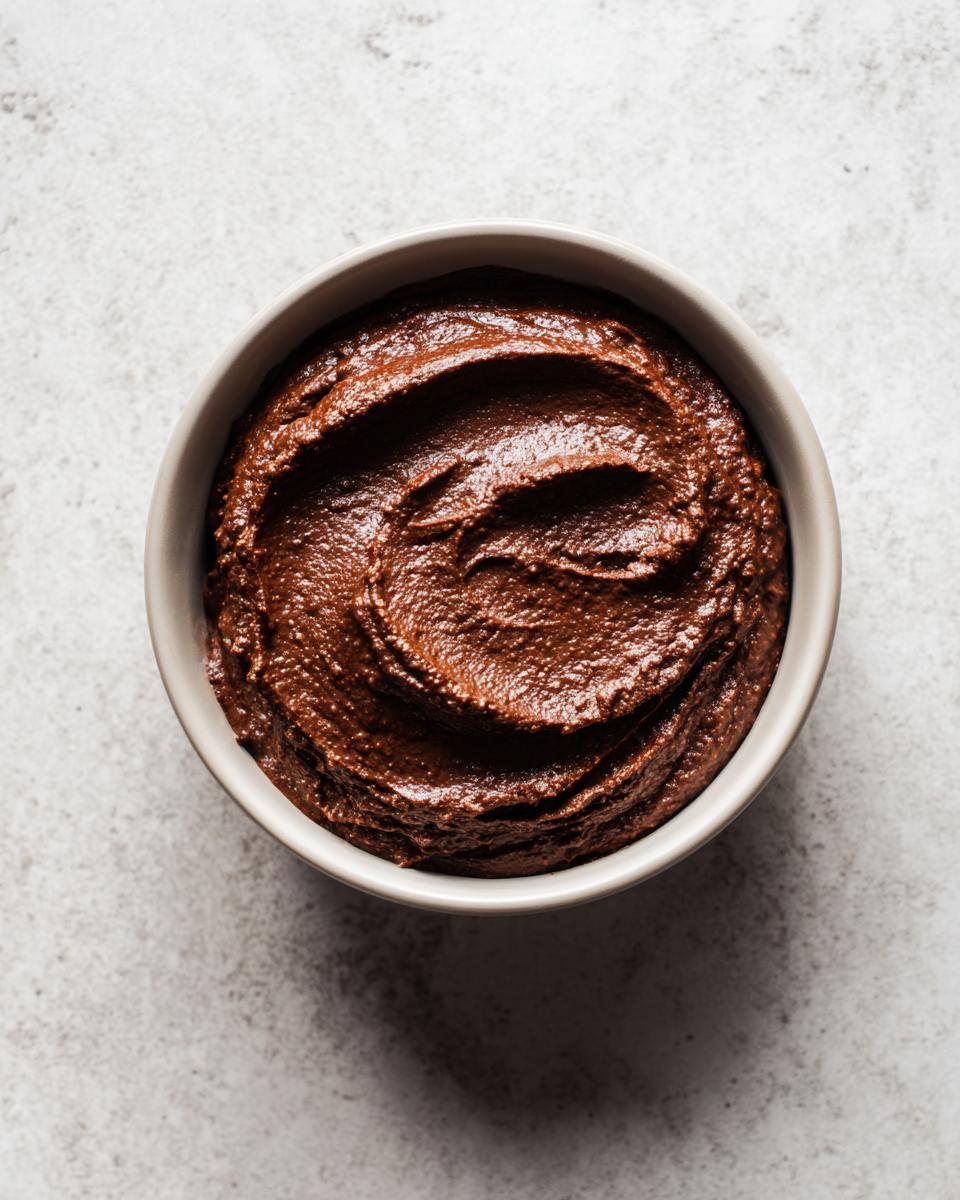 Overhead view of rich, dark chocolate dessert hummus swirled in a light gray bowl on a concrete surface.
