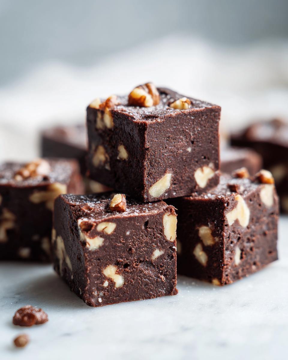 Close-up of stacked, rich chocolate walnut fudge squares sprinkled with powdered sugar on a white marble surface.