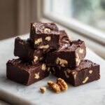 A stack of rich, dark chocolate walnut fudge squares resting on a white marble surface.