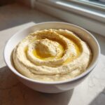 A close-up of creamy Classic homemade hummus swirled in a white bowl, drizzled with olive oil and sprinkled with spices.