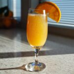 A refreshing Classic orange mimosa with bubbles, garnished with an orange slice, sitting on a countertop in sunlight.