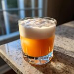 A vibrant orange Classic Whiskey Sour cocktail topped with a thick, white layer of foam, sitting on a granite counter near a window.