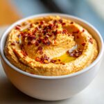 Close-up of creamy roasted red pepper hummus topped with olive oil and red pepper flakes in a white bowl.