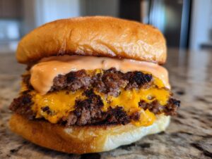 Close-up of a juicy double patty smash burger featuring crispy edges, melted cheddar cheese, and a generous drizzle of orange sauce.