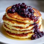A tall stack of fluffy Lemon Ricotta Pancakes topped with dark berry compote.