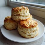 A stack of four golden-brown, flaky sourdough biscuits piled on a white plate near a window.