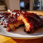 Close-up of tender, saucy Kansas City BBQ ribs glistening with dark, rich glaze under sunlight.