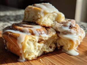 A stack of three fluffy homemade cinnamon rolls generously drizzled with thick white icing, sitting on a wooden board.