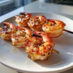 Close-up of three perfectly cooked Grilled shrimp skewers seasoned with herbs and spices, resting on a white plate.