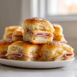 A stack of warm, glazed ham and cheese sliders topped with poppy seeds piled on a white plate.