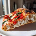 A close-up cross-section of a slice of Margherita pizza showing the airy, bubbly crust, bright red sauce, melted mozzarella, and fresh basil.