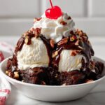 A rich nutty fudge brownie sundae featuring scoops of vanilla ice cream, chocolate syrup, pecans, whipped cream, and a maraschino cherry.