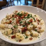 A close-up of a white bowl filled with one pan creamy gnocchi mixed with shredded chicken, topped with crispy bacon bits and fresh parsley.