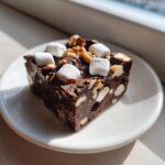 A rich square of homemade Rocky Road Fudge topped with mini marshmallows and walnuts, sitting on a white plate.