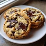 Three freshly baked soft chewy chocolate chip cookies piled on a white plate, featuring melted chocolate puddles and sea salt.